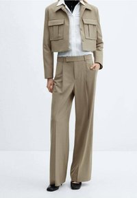 Veste beige raccourcie avec deux poches avant, associée à un pantalon large beige. En dessous, une chemise blanche à boutons. Chaussures noires.