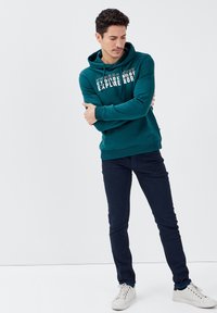 Felpa teal con testo bianco "EXPLORE MORE", dotata di cappuccio con cordini. Abbinata a jeans slim-fit blu navy e sneakers bianche.