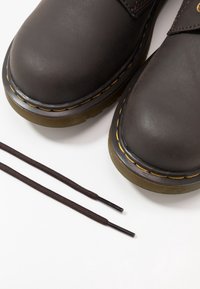 Bottes en cuir marron avec une finition lisse, surpiqûres jaunes le long de la semelle, et lacets marron reposant à côté. Elles disposent de semelles en caoutchouc robustes.