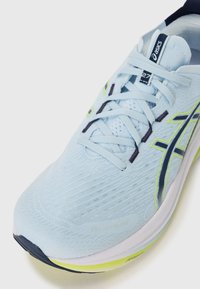 Light blue running shoe featuring a textured upper, white midsole, and black and neon green accents. Laces are slightly textured.