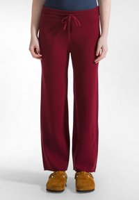 Pantalons de survêtement bordeaux avec une taille à cordon, en tissu doux, présentant une coupe décontractée et des jambes droites. Associés à des pantoufles marron.