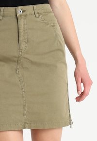 Jupe en coton kaki avec une coupe ajustée, poches avant et détail de fermeture éclair sur le côté. Texture lisse avec des accents de coutures subtiles.