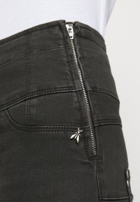 Pantalones de mezclilla negros con una cintura alta, detalle de cremallera plateada y un pequeño acento metálico en forma de abeja en el bolsillo delantero, con una textura suave.