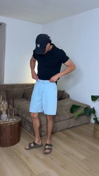 Άνδρας που φοράει μαύρο καπέλο, μαύρο πουκάμισο και ανοιχτό μπλε σορτς προσαρμόζει τα σορτς σε καθιστικό με καφέ καναπέ και πράσινο φυτό.