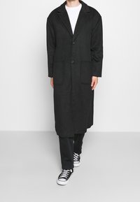 Persona che indossa un lungo cappotto nero, pantaloni neri e sneakers bianche e nere, in piedi contro uno sfondo bianco uniforme.