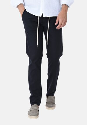 Uomo che indossa pantaloni neri con coulisse, camicia bianca e scarpe slip-on grigie, in piedi con una mano in tasca su sfondo bianco.