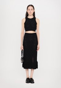 Fekete kötött cropped top és passzoló szoknya, fodros szegéllyel, oldalsó hasítékkal. Fekete cipőkkel és egy kisméretű kézitáskával viselve.