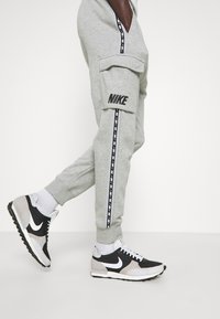 Šedé Nike tepláky se bočními kapsami a černými pruhy, na kterých je logo. V kombinaci s černými a šedými Nike teniskami.
