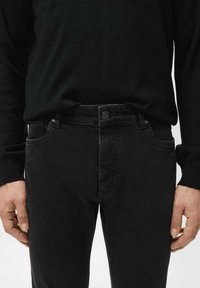 Jeans de mezclilla negros con un diseño clásico de cinco bolsillos, cierre de botones y un ligero desgaste en la parte delantera. Combinados con un suéter negro.