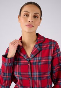 Camisa de pijama de cuadros rojos con cuello, que presenta líneas azules y blancas, detalles de botones y mangas largas. Tejido suave con un corte relajado.