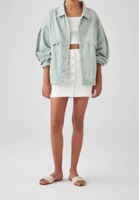 Veste en denim oversize vert clair avec des épaules tombantes, deux poches poitrine, portée sur un crop top blanc et une jupe blanche à boutons. Sandales tressées.