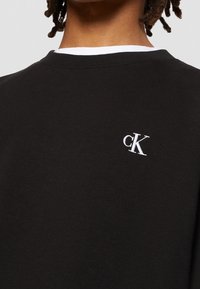 Sudadera negra de algodón, con cuello redondo, cuello acanalado y un pequeño logo blanco "cK" en el pecho. Textura suave.