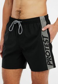 Zwarte zwembroek met een elastische tailleband en een trekkoord. Beschikt over een zijstreep met het woord "PROTEST" in het wit, gladde textuur.