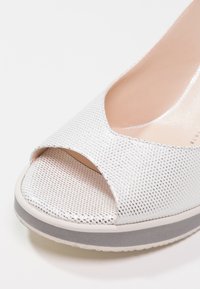 Chaussure blanche à bout ouvert avec une surface supérieure perforée, une doublure beige douce et une semelle en caoutchouc grise. Présente un motif lisse et texturé.