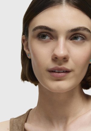 Pendientes de botón en tono azul verdoso con una forma geométrica, lucidos sobre un cabello corto castaño. Los pendientes destacan sobre un tono de piel neutro y un fondo también neutro.