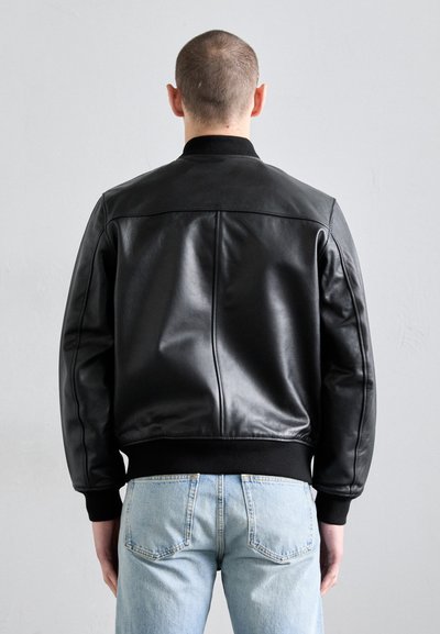 Svart läder bomberjacka med räfflad krage och manschetter, med en slät yta och en strukturerad design. Bärs över ljusblå jeans.