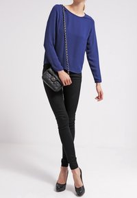 Nainen, joka pukeutunut pitkähihainen sininen paita, mustat kapealahkeiset housut, mustat korkokengät ja kantaa mustaa tikattua olkalaukkua ketjuhihnalla.