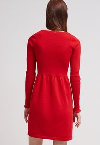 Abito rosso a maniche lunghe con corpino aderente e gonna svasata. Tessuto morbido con una consistenza liscia, caratterizzato da un colletto rotondo e dettagli delle cuciture.