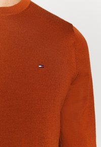 Gros plan sur un pull en maille orange rouille avec un petit patch logo rectangulaire sur la poitrine gauche, comprenant des couleurs rouge, blanc et bleu marine.