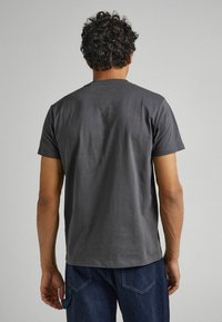 Hombre con cabello rizado que lleva una camiseta básica de manga corta gris oscuro y jeans azul oscuro, de pie de espaldas contra un fondo neutral.