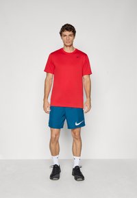 Young man standing, wearing a red Nike t-shirt, blue Nike shorts, white Nike socks, and black athletic sneakers against a plain background.