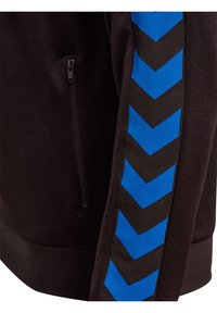 Sort trøje med lynlås-lomme på siden, prydet med blå og sorte chevron-striber langs ærmet, fremstillet af glat stof.