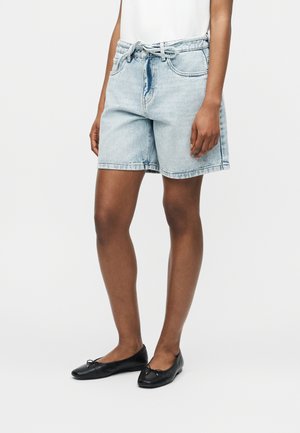 Modelka nosząca jasnoniebieskie jeansowe szorty z przewiązanym paskiem, biały top oraz czarne baleriny, stojąca na jednolitym białym tle.