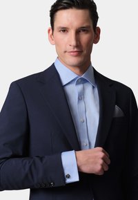 Navy blazer with a light blue shirt, featuring a textured fabric, silver cufflinks, and a patterned pocket square. Close-up on top half of model.