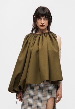 Nainen pukeutuneena epäsymmetriseen oliivinvihreään kokoontaitettuun toppiin, jossa on yksi hiha, yhdistettynä korkealaiseen ruudulliseen hameeseen, seisoo yksivärisen taustan edessä.