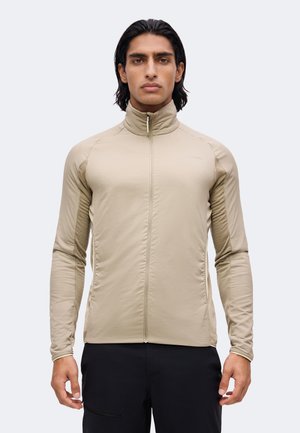 Mann mit mittellangen dunklen Haaren, der eine beige Jacke mit Reißverschluss und schwarze Hose trägt, steht vor einem schlichten hellen Hintergrund.