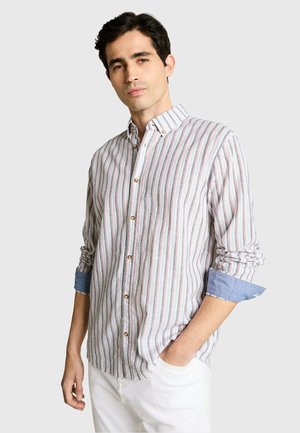 Man draagt een overhemd met lange mouwen, verticale strepen en opgerolde blauwe manchetten, gecombineerd met een witte broek, staand met één hand in zijn zak.