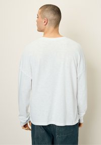 Ilgas rankovėmis marškiniai iš lengvos, tekstūruotos baltos medžiagos, su laisvu siluetu ir nukirptais pečiais, bei apvalia briauna ties juosmeniu.