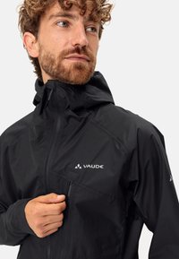 Giacca impermeabile nera con cappuccio, dotata di tasca con zip e accenti di logo riflettente. Materiale leggero e design elegante.