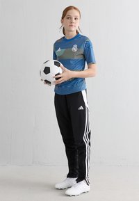 Jauns cilvēks zilajā Real Madrid krekla un melnās Adidas sporta biksēs, turot melnbalto futbola bumbu, stāv uz pelēka grīdas.