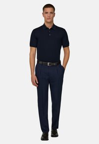 Jeune homme debout, les mains dans les poches, portant un polo bleu marine, un pantalon à carreaux bleu marine, une ceinture noire et des chaussures noires.