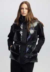 Chaleco puffer negro con un acabado brillante, que presenta un cuello alto, cierre de botones, cintura elástica y bolsillos, superpuesto a una camiseta negra.