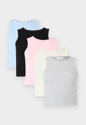 Jeune femme aux cheveux bouclés portant un crop top blanc sans manches, un pantalon large noir et des baskets noires, debout sur un fond blanc.