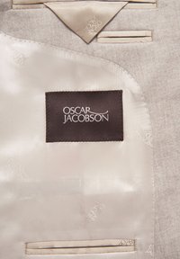 Ljust grå kostyminredning med krämfärgad satinfoder, med en mörkbrun etikett där "OSCAR JACOBSON" är broderat i vitt. Slät textur.