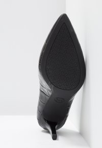 Chaussure à talon haut noire à bout pointu, inclinée pour montrer la semelle texturée avec un motif répétitif du logo "MK" et un petit logo circulaire près du talon.