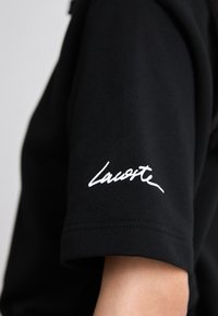 Čierne tričko s krátkym rukávom s bielym písaným logom "Lacoste" na ramene, nosené na hornom ramene osoby.