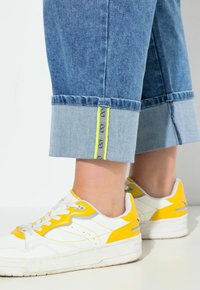 Pantalones anchos de mezclilla azul claro con puños doblados, con un acento amarillo cosido a lo largo del dobladillo. Combinados con zapatillas blancas destacadas en amarillo.