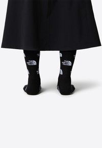 Svarta strumpor med "THE NORTH FACE"-logotyp i vitt, med en texturerad yta och kontrasterande band. Bärs tillsammans med en lång svart kjol.