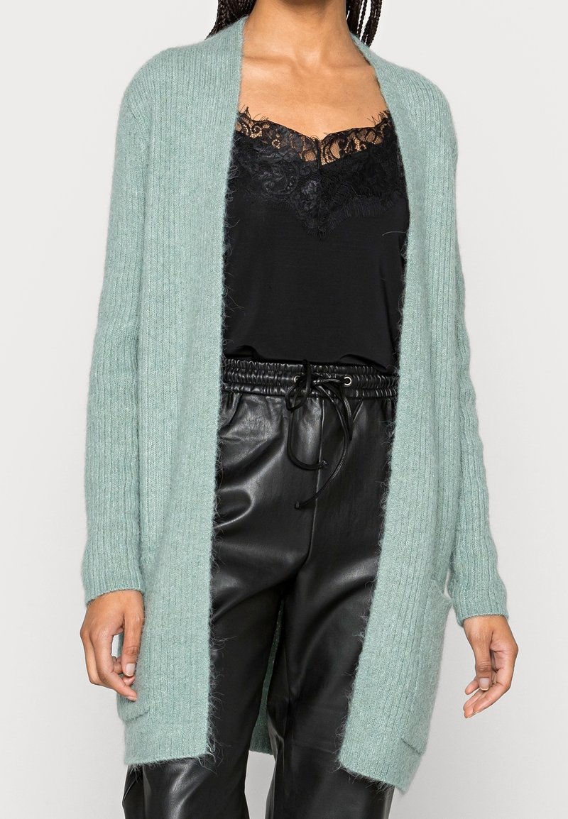 Femme portant un cardigan côtelé vert clair sur un haut noir orné de dentelle, avec un pantalon en cuir noir taille haute noué par un cordon.