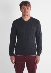 Pull en maille gris foncé avec un col en V, des manches longues et une bordure contrastante gris clair au bas. Porté avec un pantalon bordeaux.