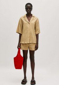 Beige Kurzarmhemd mit strukturiertem Muster und Kragen, kombiniert mit passenden Shorts. Hält eine rote strukturierte Tote-Tasche. Schwarze Sandalen.