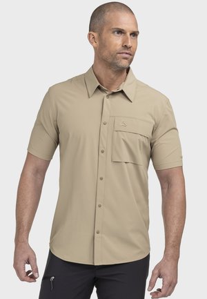 Mann trägt ein beige kurzärmeliges Hemd mit Brusttasche mit Klappe und schwarze Hose, steht vor einem schlichten Hintergrund.