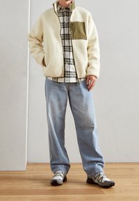 Persoon draagt een crèmekleurig fleecejack met een olijfgroene borstzak, een geruit overhemd, lichtblauwe spijkerbroek en grijze wandelschoenen, staande op een houten vloer.