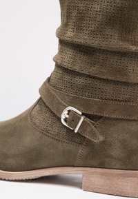 Bottine en daim vert avec un shaft texturé, une lanière décorative à boucle et un talon en bois. Présente une finition douce et lisse.