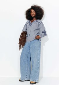 Blouse grise avec des accents brodés, large jean en denim clair, sac à franges en suède marron, bottines marron, tissu texturé, coupe décontractée.