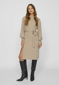 Vestito a maglia beige con maniche lunghe, cintura in vita e spacco laterale; abbinato a stivali neri in pelle al ginocchio. Tessuto liscio e design aderente.